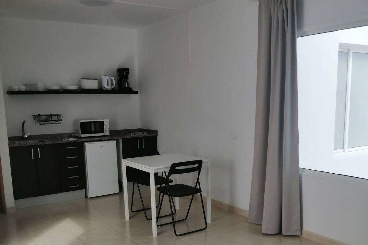 A compact kitchen with a microwave, toaster, and coffee maker on a black countertop. A small refrigerator is below the countertop. There is a dining table with two chairs. The room has a window with curtains and a tiled floor.