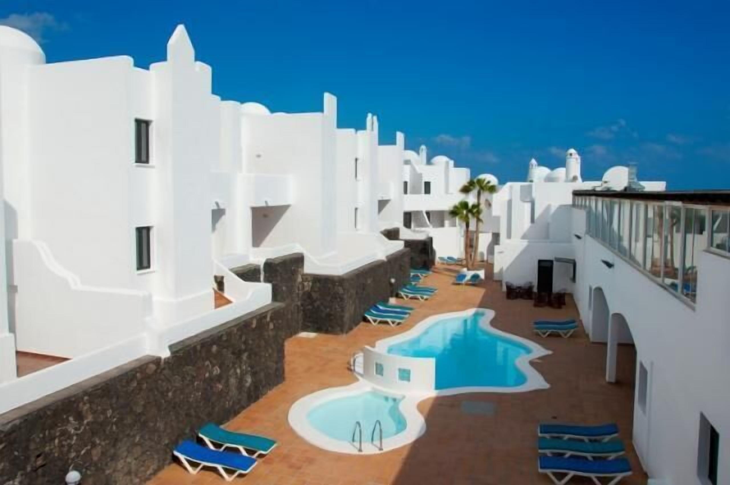 Zona de piscina en la azotea con edificios blancos, sillones azules y una piscina.