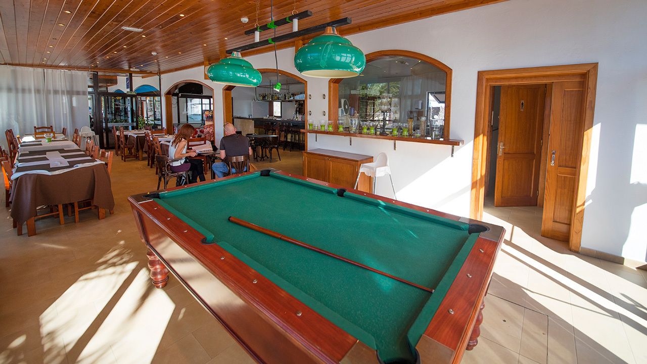 Una mesa de billar en una sala con techos de madera y ventanas amplias. Hay mesas y sillas alrededor de la mesa de billar, y una zona de bar al fondo.