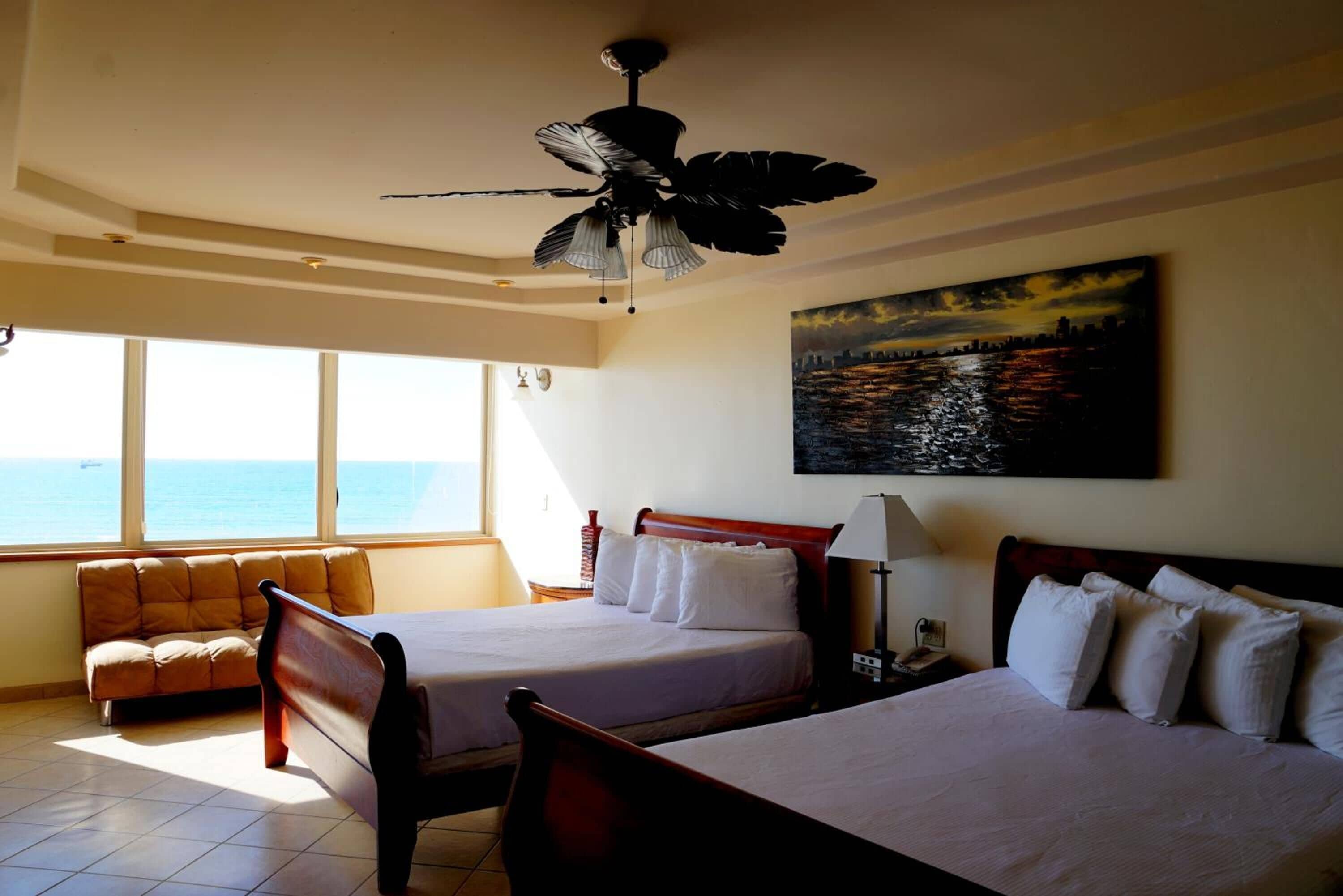Een hotelkamer met twee bedden, een slaapbank, een plafondventilator, een schilderij van een stadsgezicht en uitzicht op de oceaan.