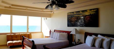 A hotel room with two beds, a sofa, a ceiling fan, a painting of a cityscape, and a view of the ocean.