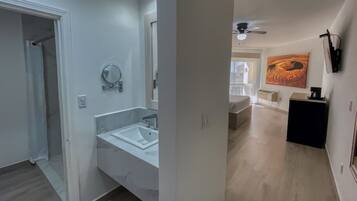 Un baño moderno con un lavabo y espejo blancos, zona de ducha y pasillo que conduce a una sala de estar con sofá y obras de arte.
