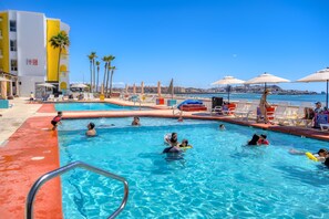 A swimming pool with people, palm trees, and a building with yellow and white colors. - Playa Bonita (Puerto Peñasco)