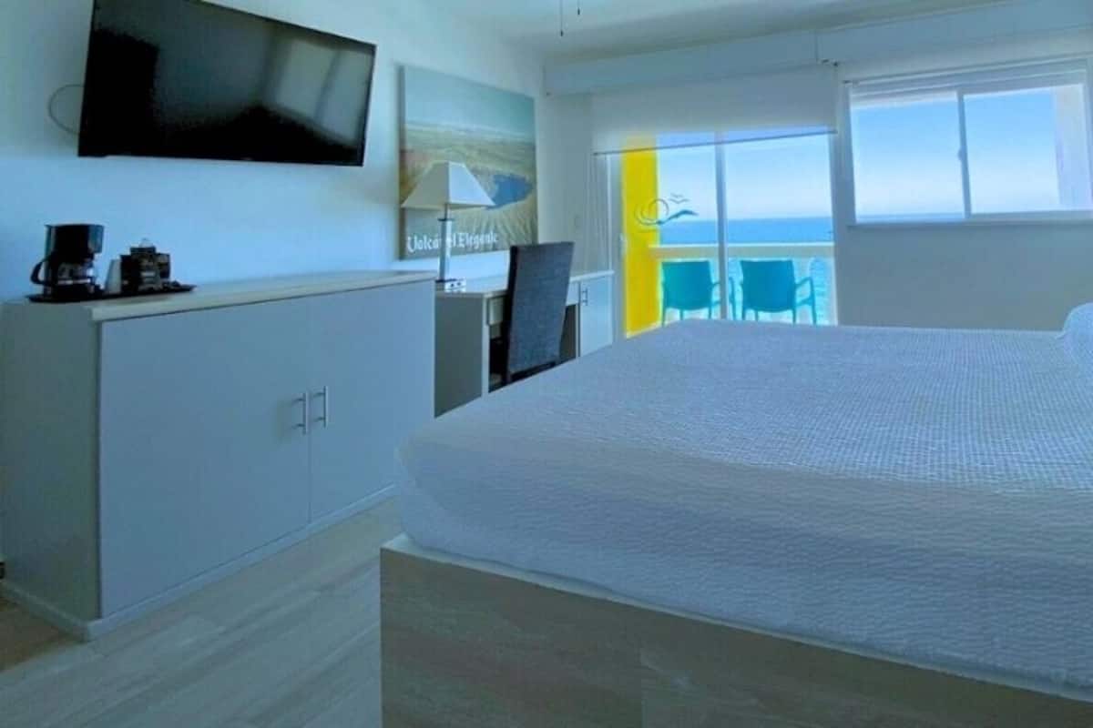 Une chambre d’hôtel avec un lit, une télévision, un bureau avec une lampe et une vue sur la mer depuis un balcon.