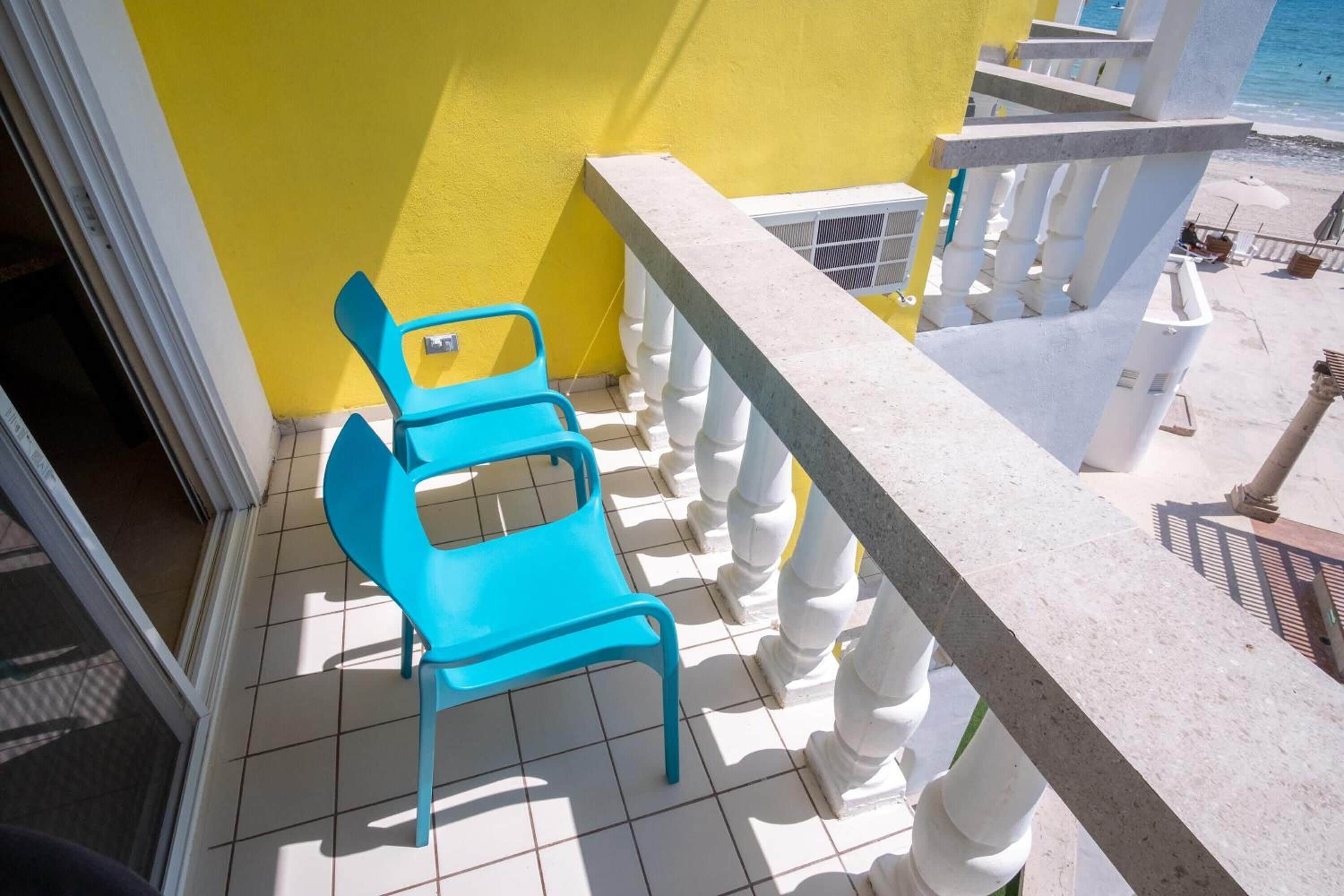 Un balcón con una pared amarilla, barandilla blanca y un par de sillas azules.