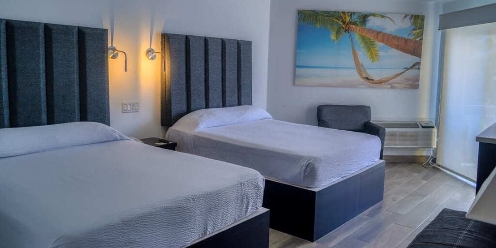 A hotel room with two beds, a painting of a beach, and a window with curtains.