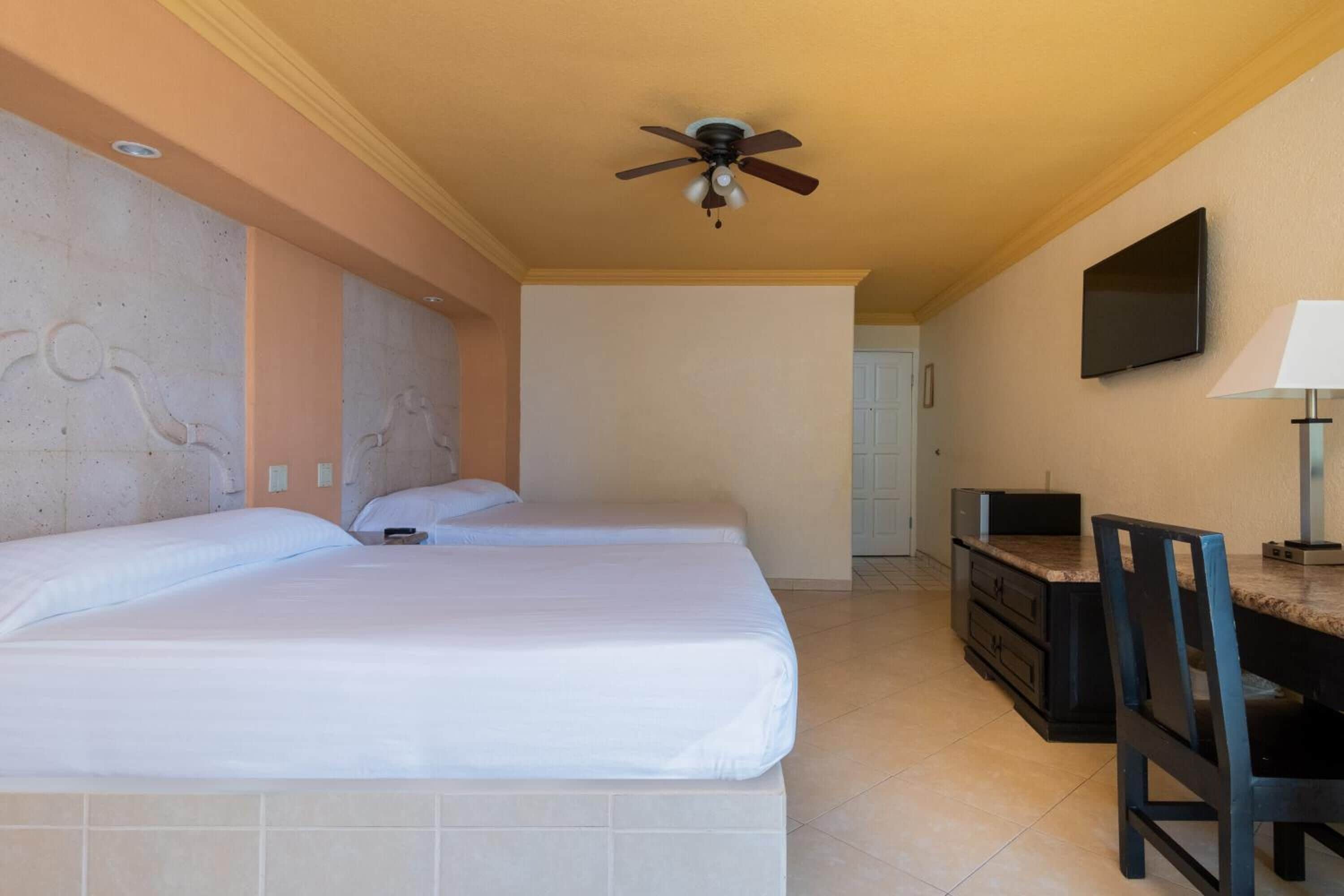 A hotel room with two beds, a ceiling fan, a television mounted on the wall, and a desk with a chair.