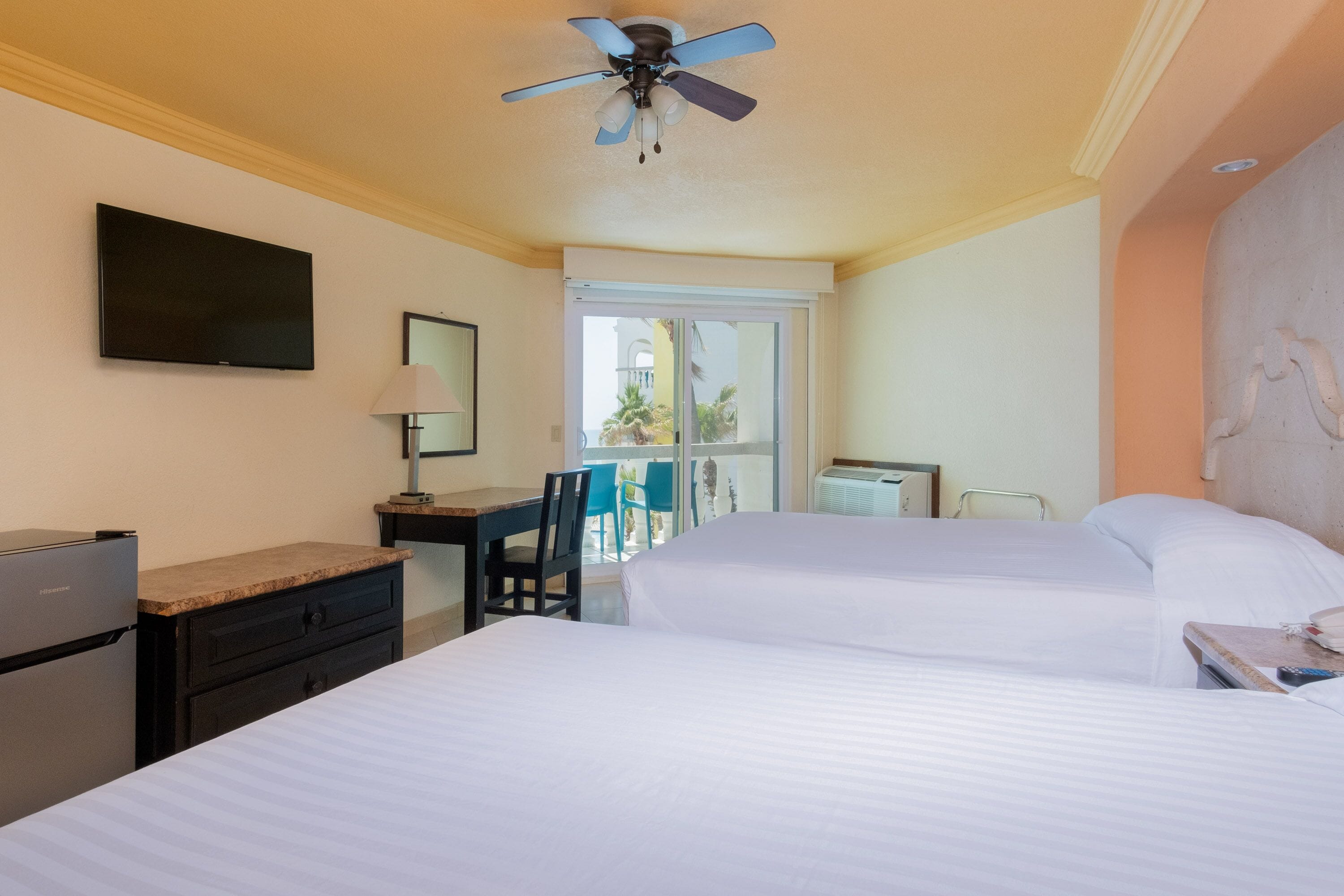 A hotel room with two beds, a desk, a chair, a television, and a ceiling fan.