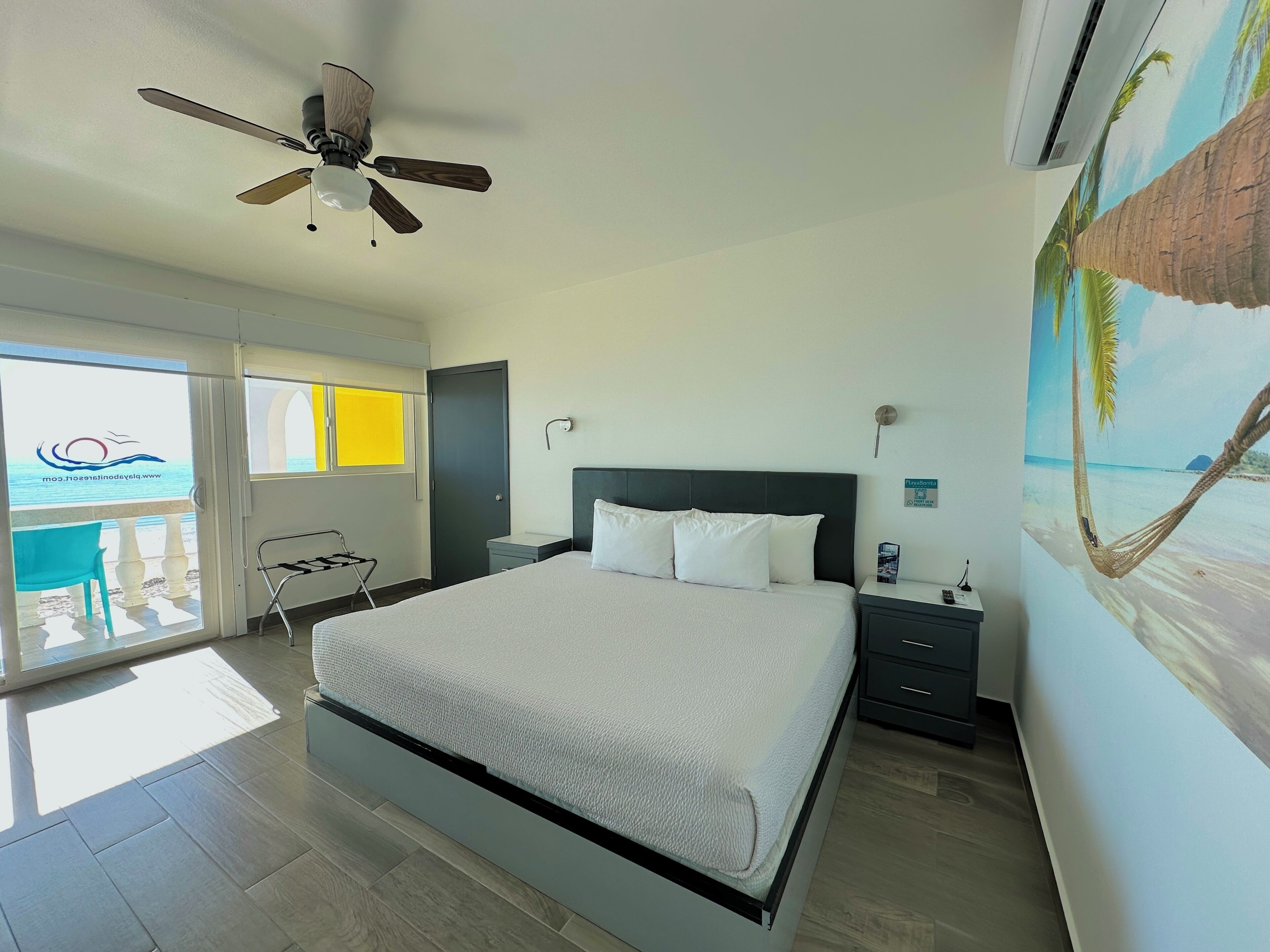 A bedroom with a bed, a ceiling fan, a nightstand, a wall-mounted air conditioner, and a beach-themed mural.