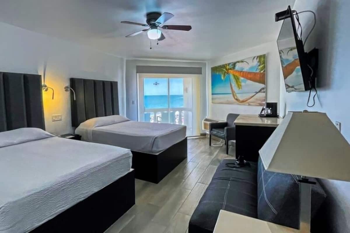 Une chambre d’hôtel avec deux lits, un bureau, une chaise, un ventilateur de plafond, une télévision et un tableau représentant un palmier.