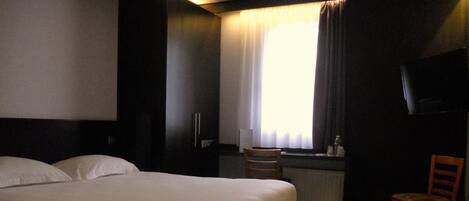 Quarto de hotel com uma cama grande, uma escrivaninha, uma cadeira, uma televisão e uma janela com cortinas.