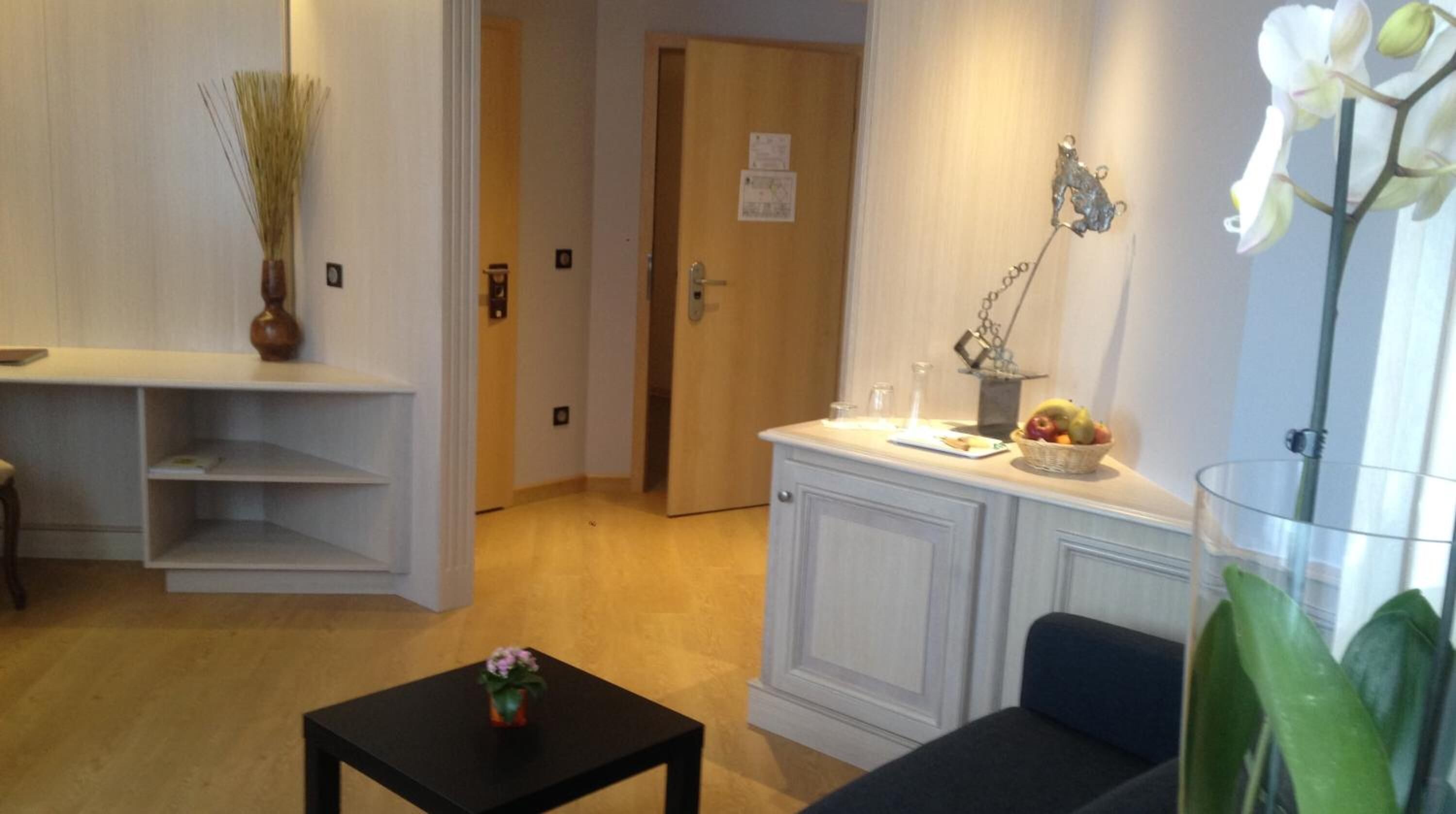 A room with a wooden floor, a white door, a small table with a vase, a blue sofa, and a wall-mounted shelf with a plant.