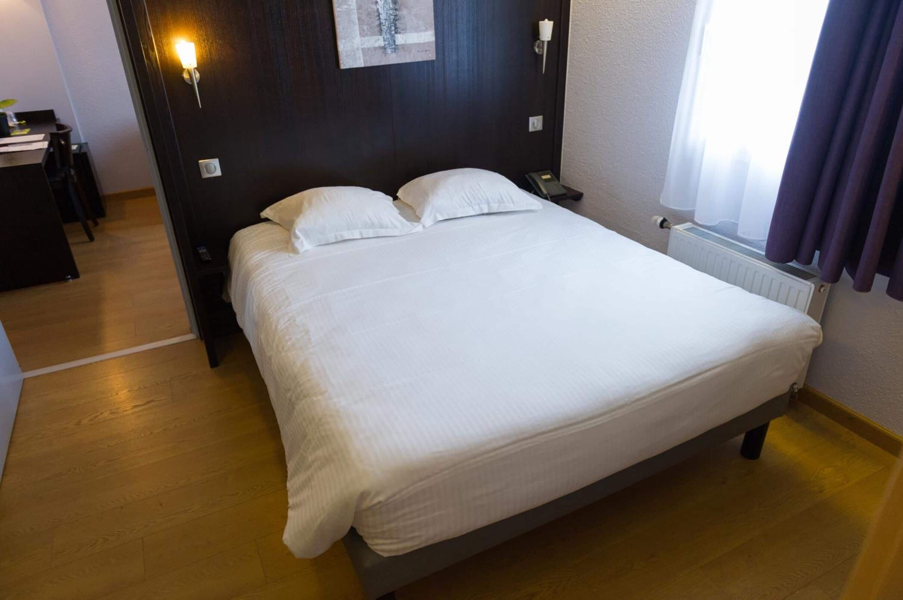 Una cama bien hecha en una habitación de hotel, con suelo de madera, una mesita de noche con lámpara y una ventana con cortinas.