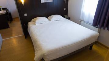 Una cama bien hecha en una habitación de hotel, con suelo de madera, una mesita de noche con lámpara y una ventana con cortinas.