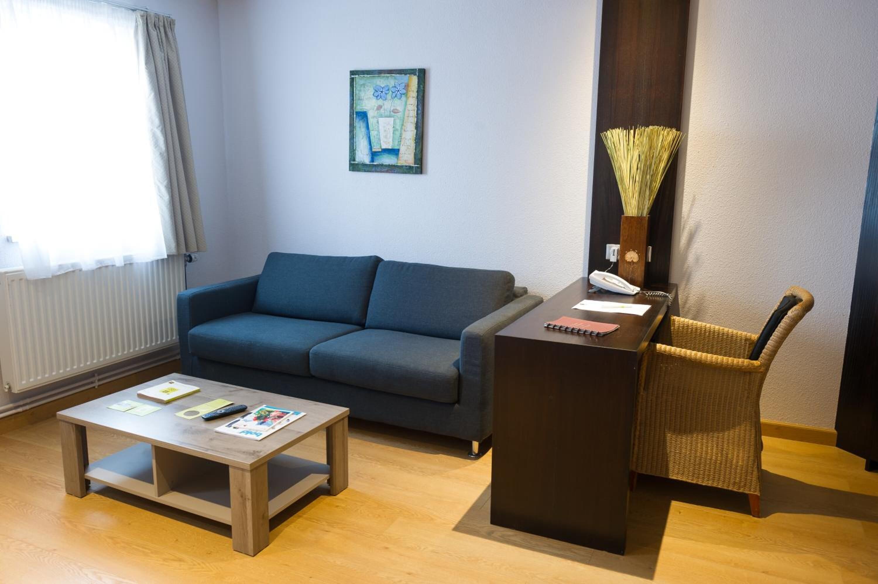 Una sala de estar con un sofá azul, una mesa de centro de madera, un escritorio oscuro con un jarrón de plantas secas y una silla de mimbre.