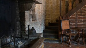 A rustic room with a stone wall, a wooden chair, a fireplace with a metal grate, and a staircase with wooden railings. - Auberge Le Renard'eau (Beze)