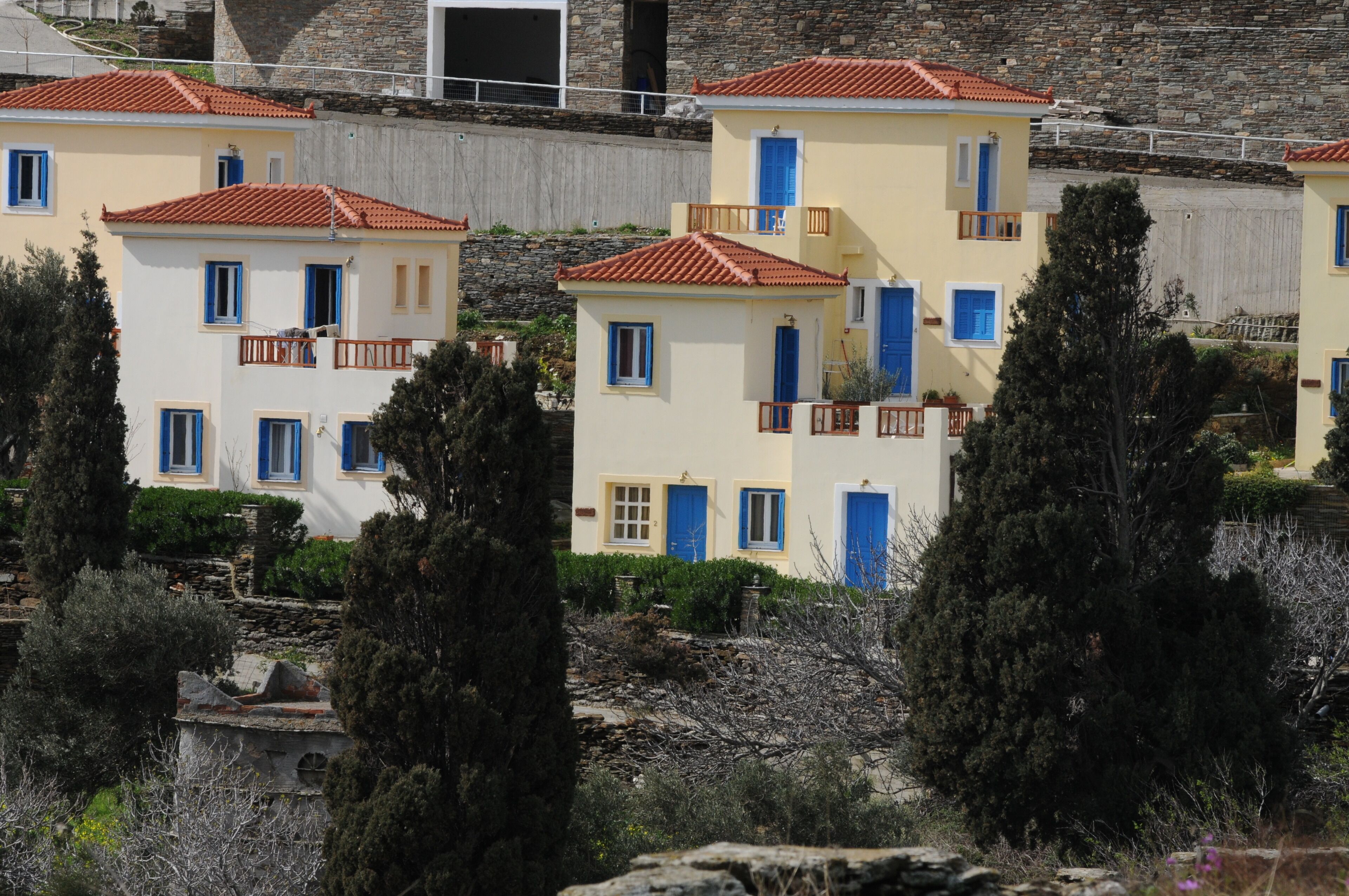 Une rangée de bâtiments de style méditerranéen, avec des toits couverts de tuiles rouges et des portes bleues, entourés d’une végétation luxuriante et d’arbres.