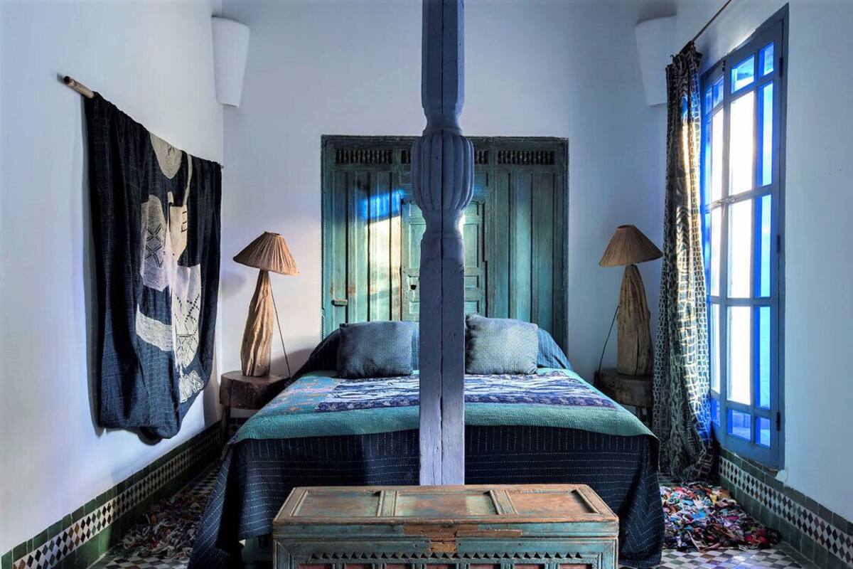 A traditional room with a bed, two lamps, a wooden chest, and a window with blue shutters.