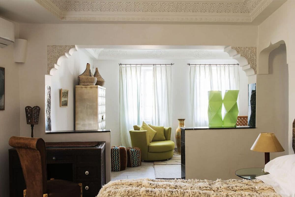A room with a bed, a desk, a chair, and a large mirror. There are curtains on the windows and a lamp on the desk.