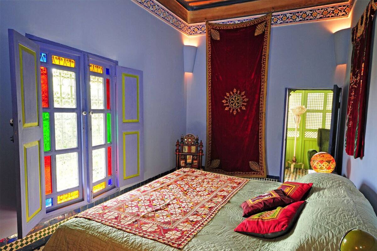 A room with a bed, colorful stained glass windows, a red curtain, and a patterned rug.