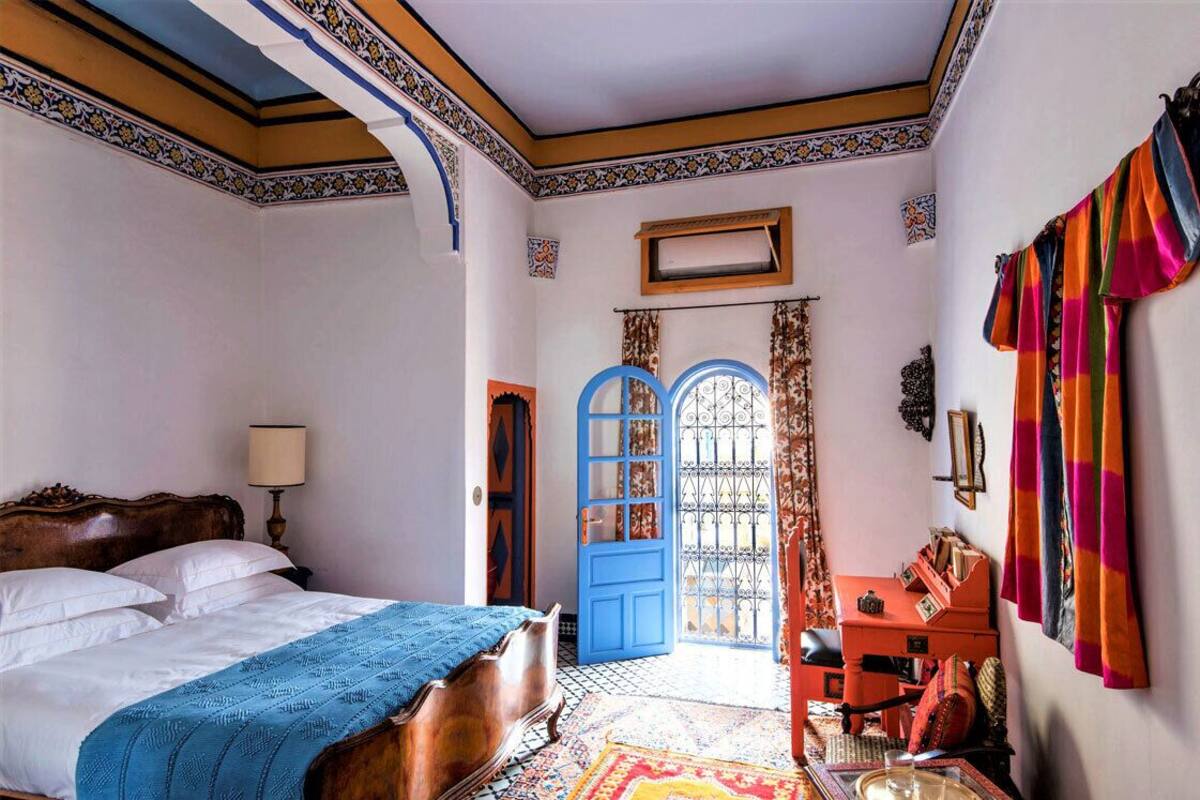 A bedroom with a bed, a desk, and a chair. There is a blue door and a window with curtains.
