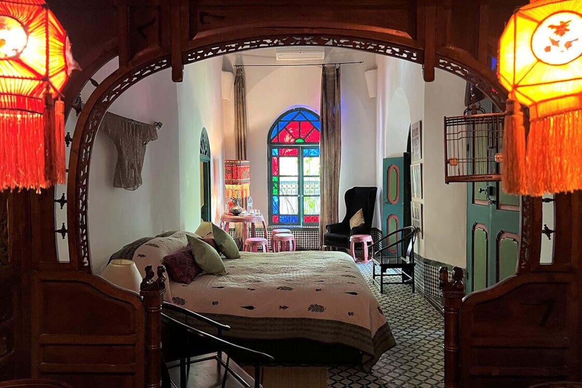 A bedroom with a bed, a chair, a table, and a window with stained glass.