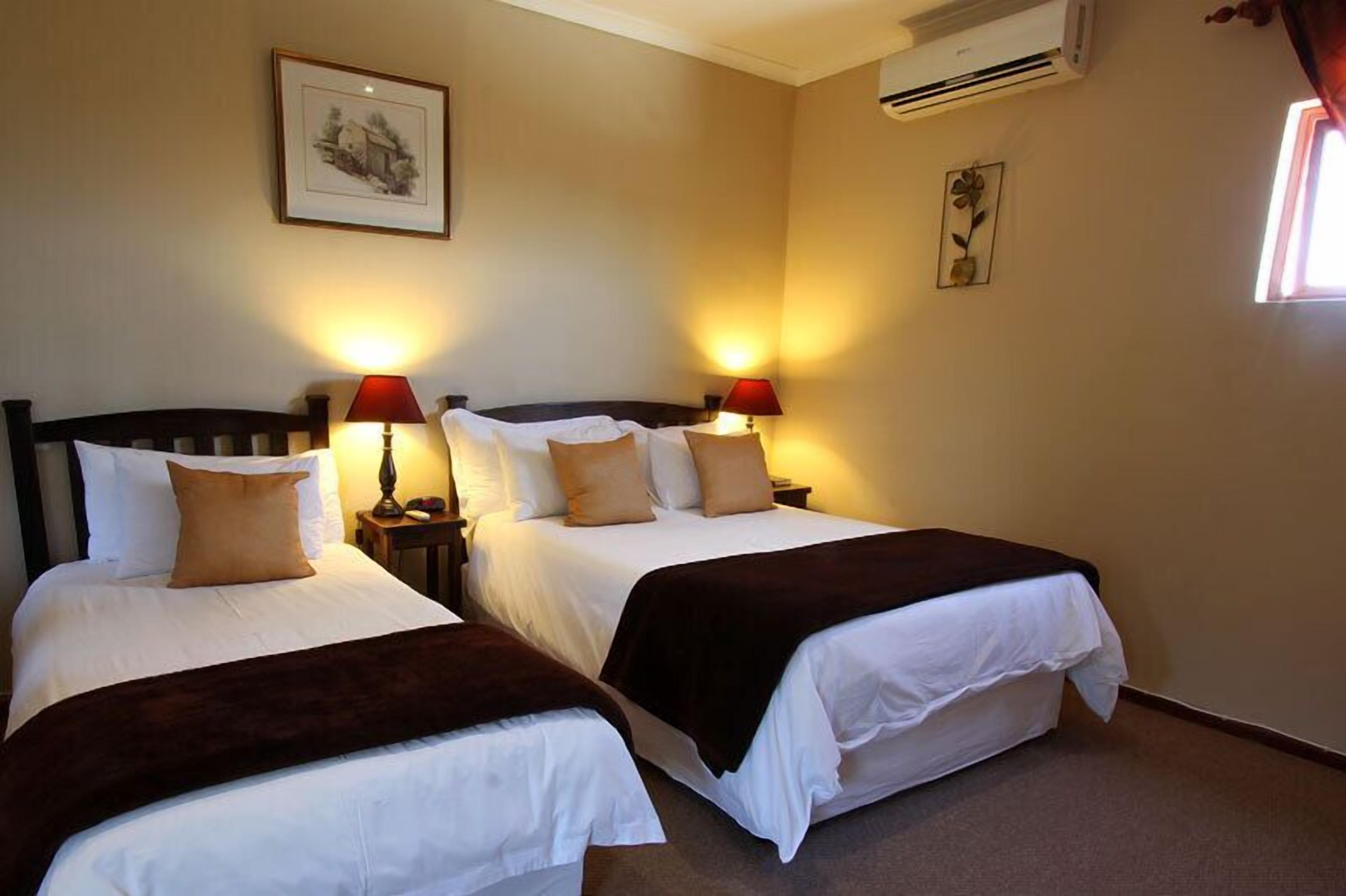 A hotel room with two beds, each with white bedding and brown throw blankets. There are two bedside lamps with red shades, and a framed picture on the wall above the beds.
