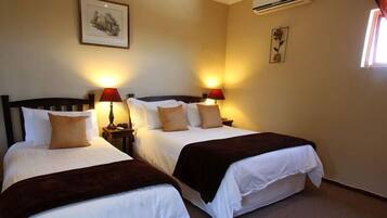 A hotel room with two beds, each with white bedding and brown throw blankets. There are two bedside lamps with red shades, and a framed picture on the wall above the beds.
