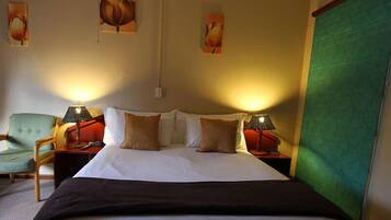 A hotel room with a large bed, two bedside tables with lamps, a green armchair, and three wall-mounted paintings.
