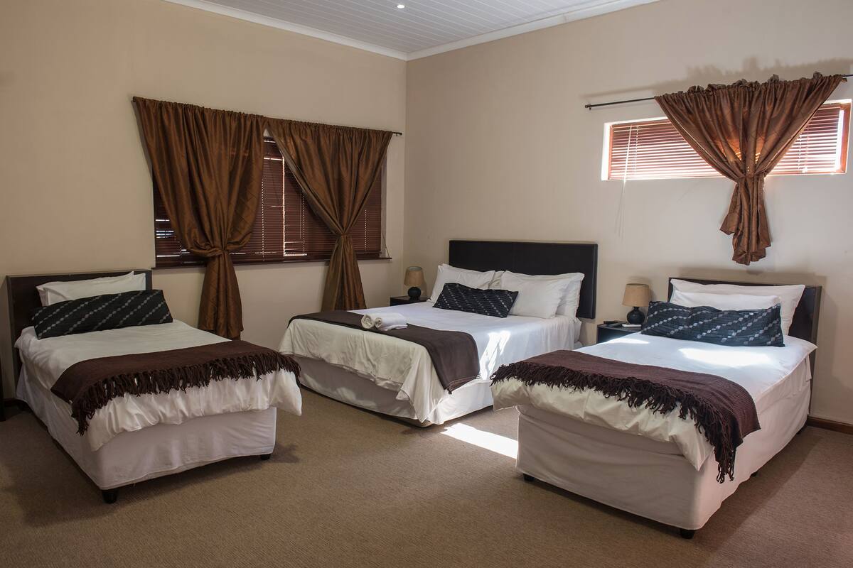 A hotel room with three single beds, each with a white mattress and brown headboard. The room has beige walls and a carpeted floor.