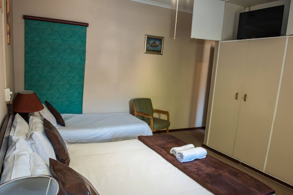 A hotel room with two beds, a green chair, a wardrobe, and a television on the wall.