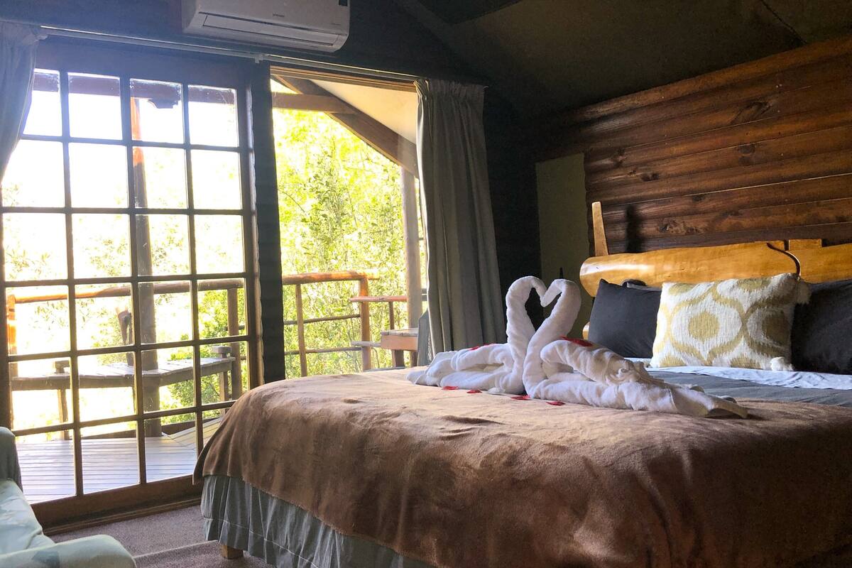 A bed with a brown bedspread, a wooden headboard, and two pillows. There is a window with curtains, and a wooden railing outside.
