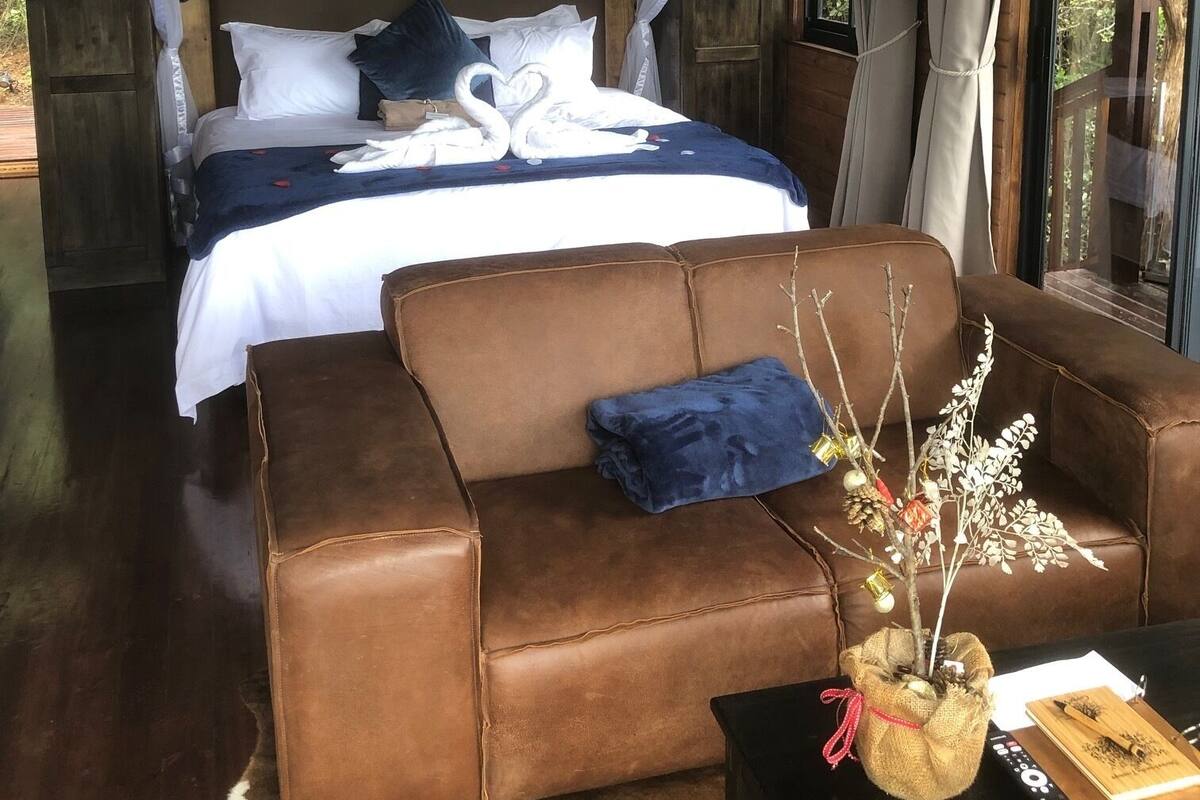 A bedroom with a bed under a canopy, a brown leather sofa, a small table with a vase and a book, and a window with curtains.