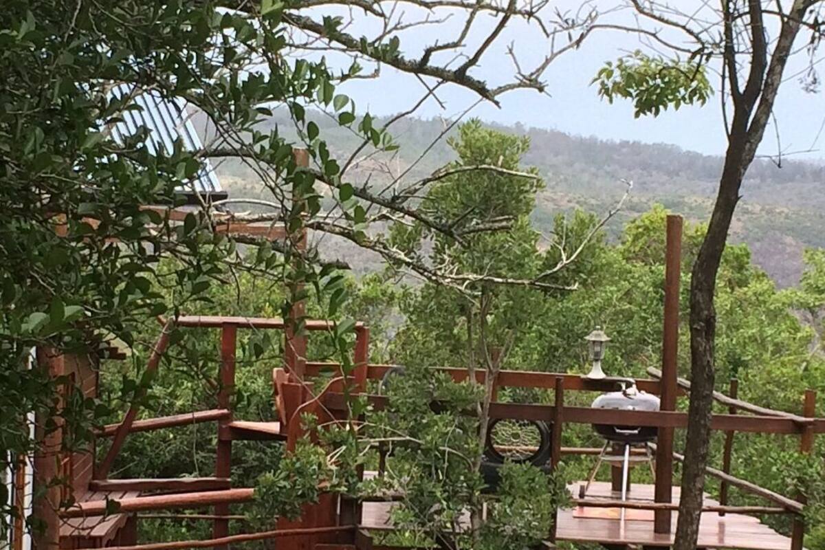 A wooden deck with a table and chairs, surrounded by greenery and a distant view of hills.