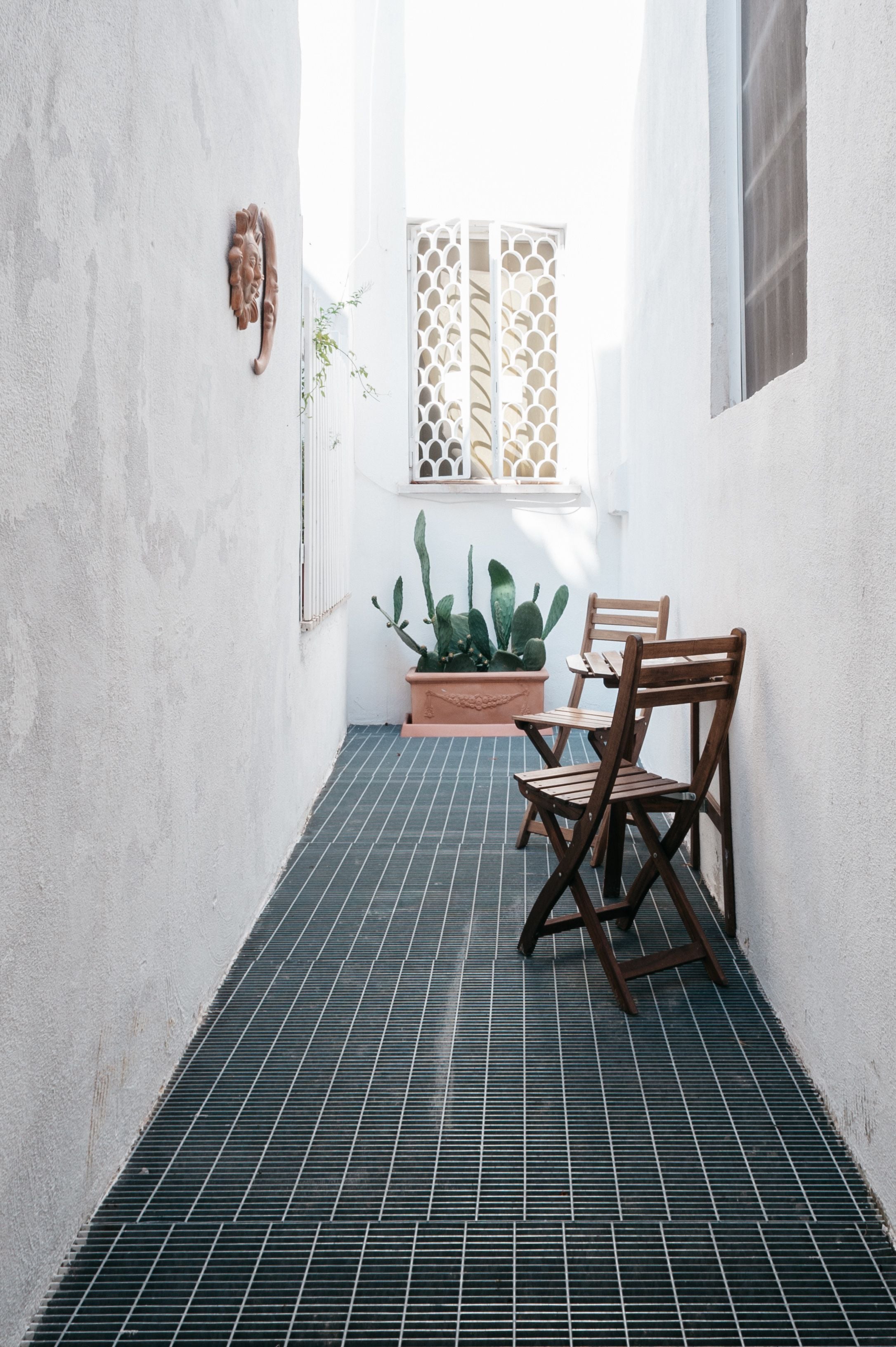 A narrow corridor with a tiled floor, two wooden chairs, a potted cactus, and a window with decorative lattice.