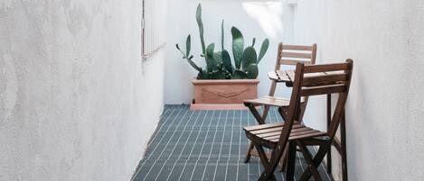 Un pasillo estrecho con suelo de baldosas, dos sillas de madera, un cactus en maceta y una ventana con celosía decorativa.