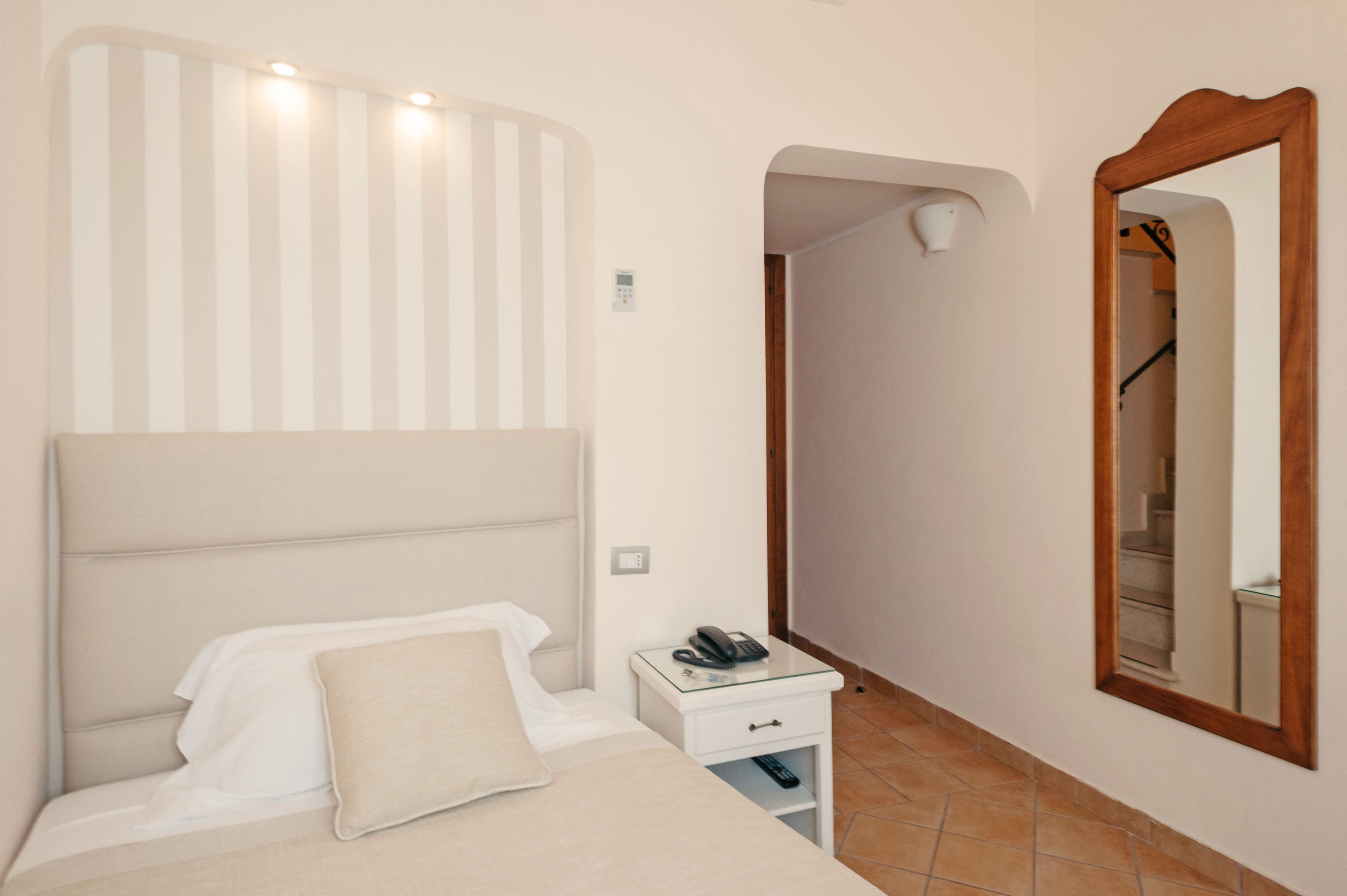 A hotel room with a bed, a bedside table with a phone, a mirror, and a wall-mounted light fixture.