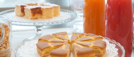 A breakfast setting with a glass plate of cake, a glass jar of granola, and two pitchers of orange and red beverages.