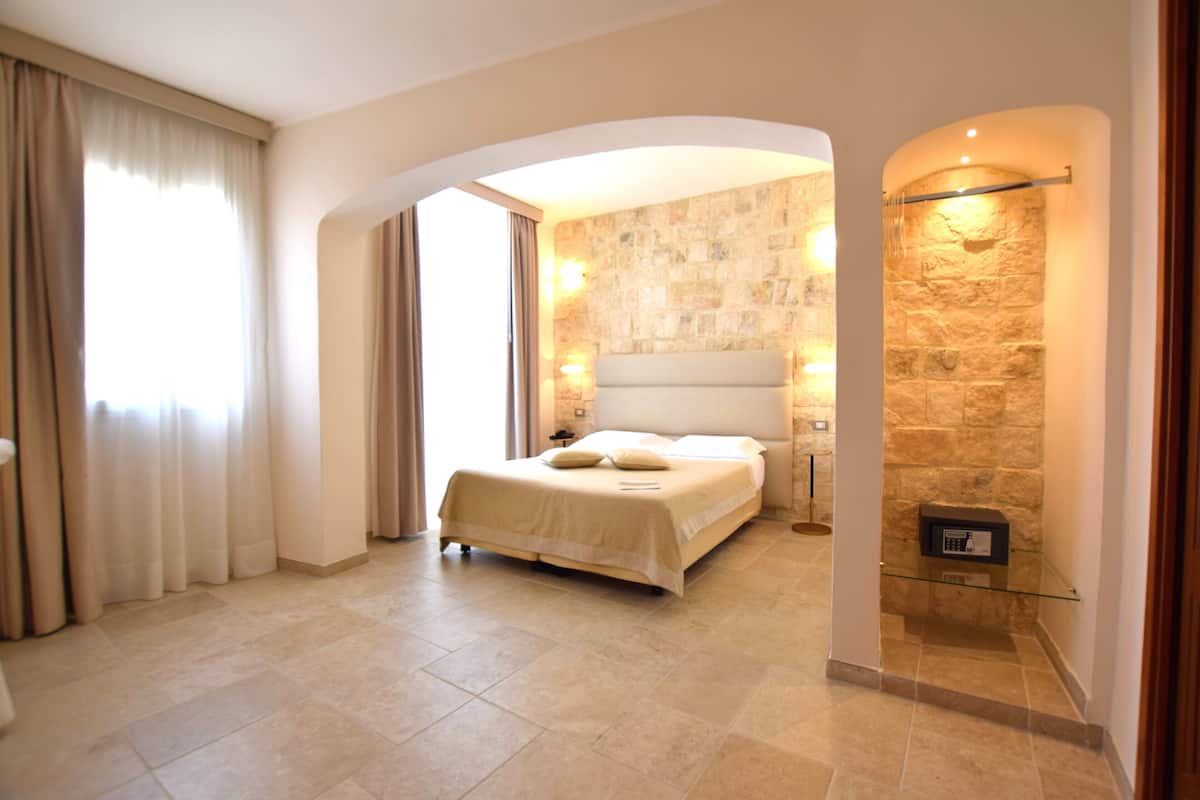 A modern hotel room with a large bed, stone walls, and a glass-enclosed shower area.