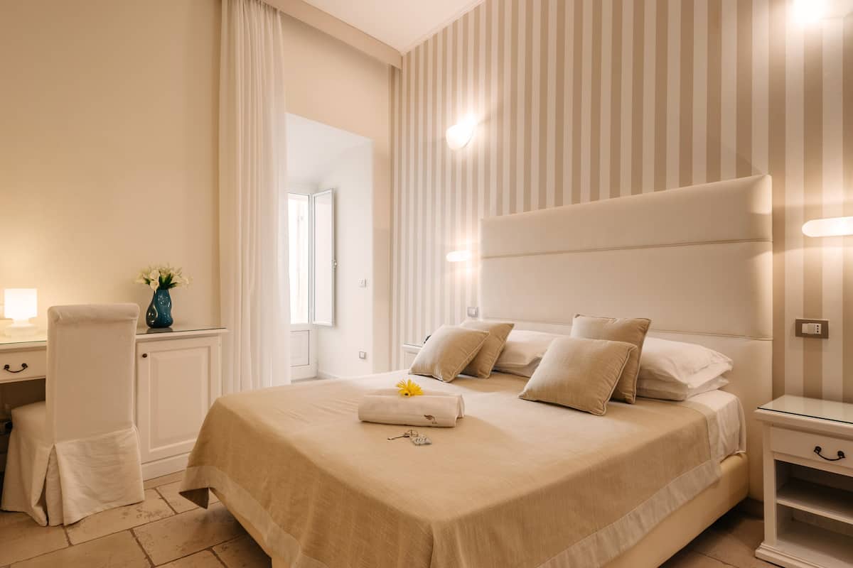 A hotel room with a large bed, a bedside table, a desk with a chair, and a window with curtains.