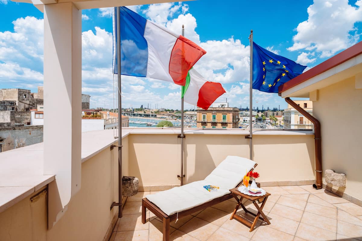 A balcony with a lounge chair and a small table, flags of Italy and the EU, and a cityscape view.