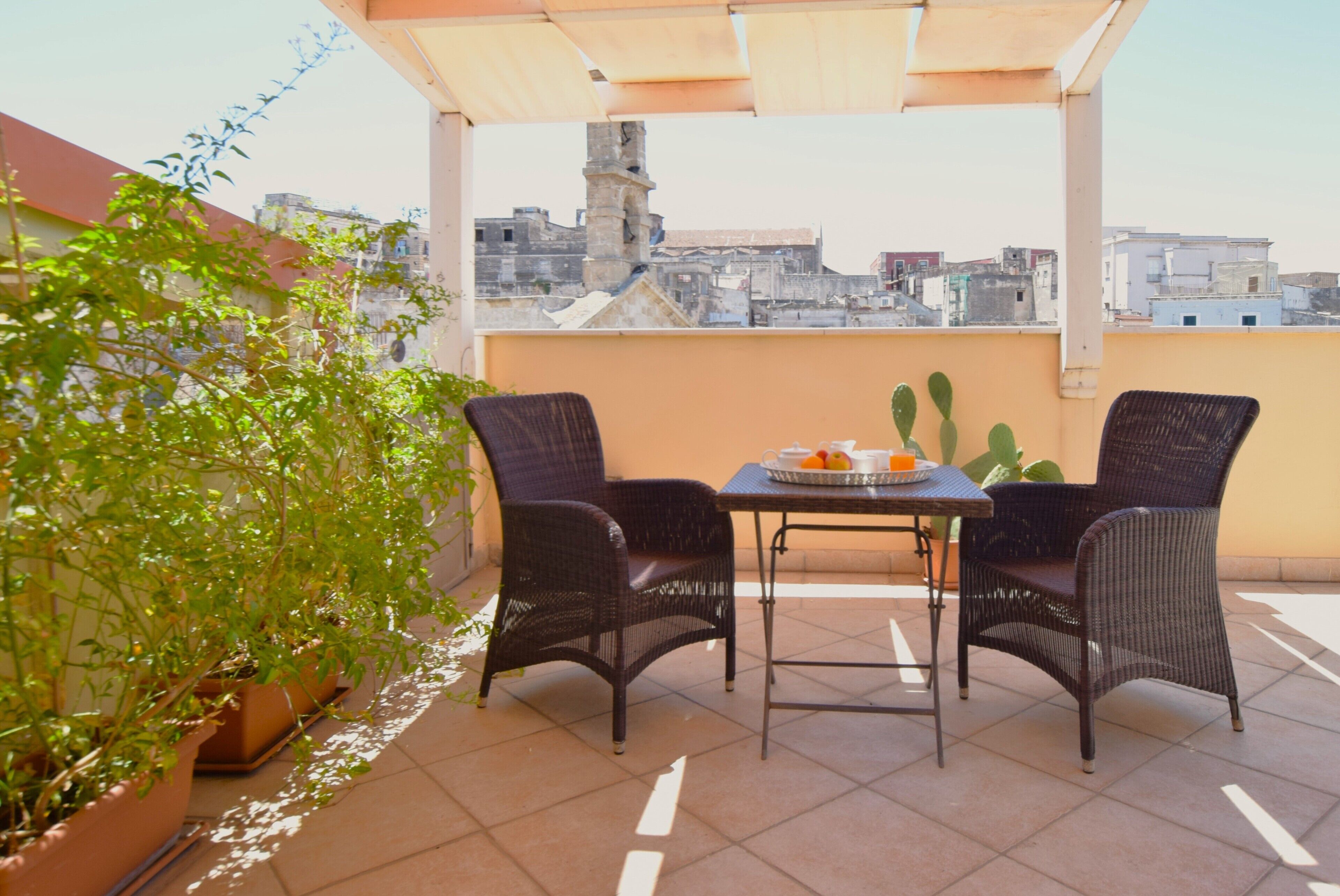 Una terraza en la azotea con dos sillones de mimbre, una mesa con una bandeja de naranjas y plantas en macetas.