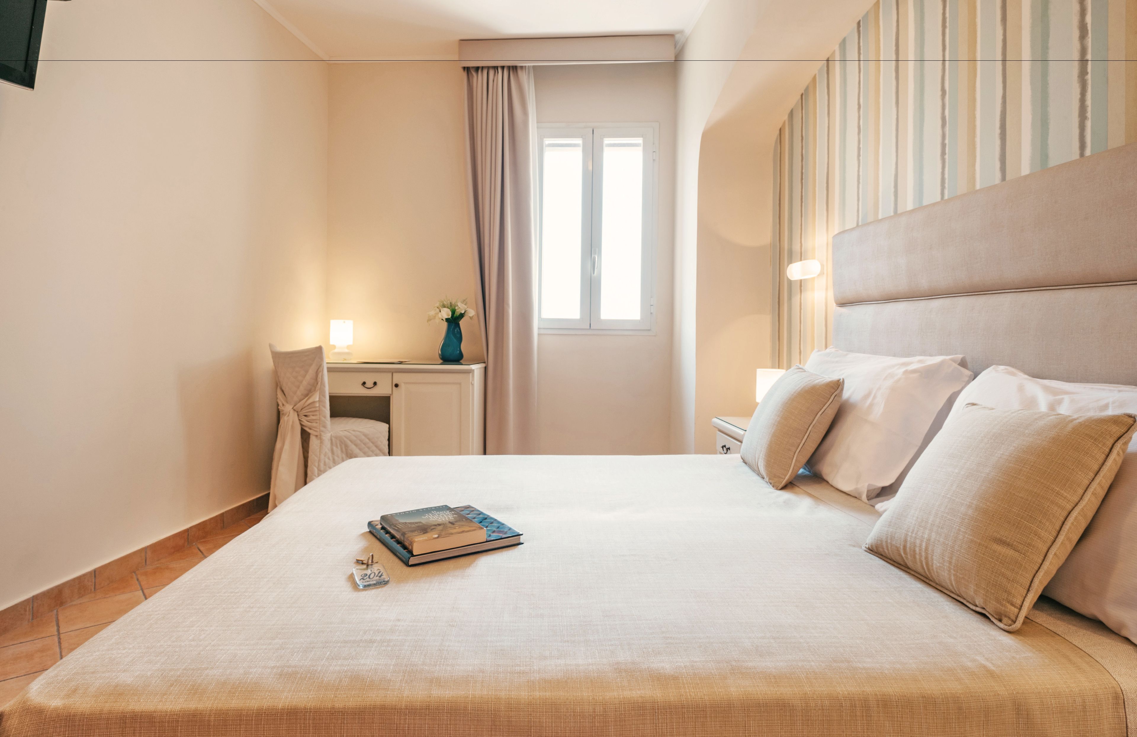 Een netjes opgemaakt bed met kussens, een nachtkastje met een lamp en een raam met gordijnen.