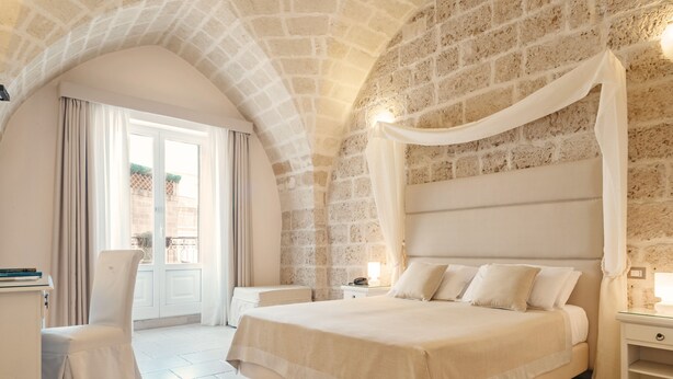 A bedroom with a stone wall, a bed with white bedding, a nightstand with a lamp, and a chair. - Hotel L'Arcangelo Boutique Hotel (Taranto)