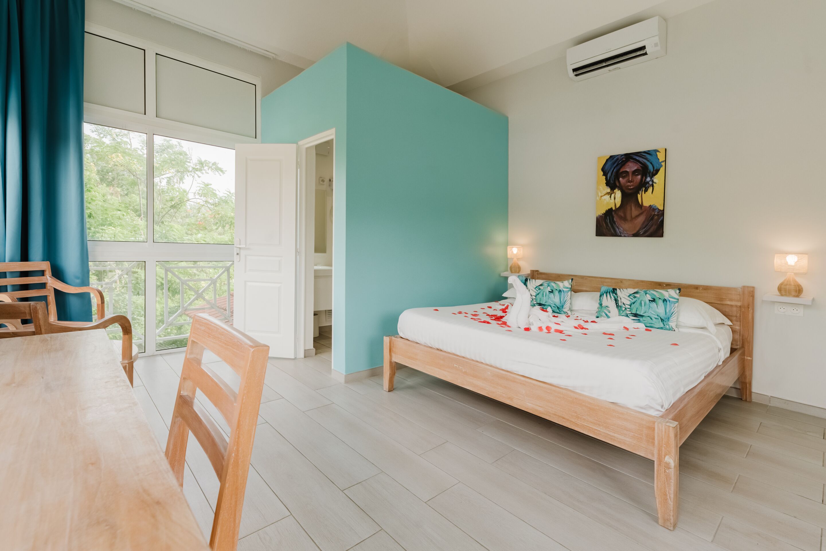 A bedroom with a wooden bed, a dining table with chairs, a large window with curtains, and a painting on the wall.