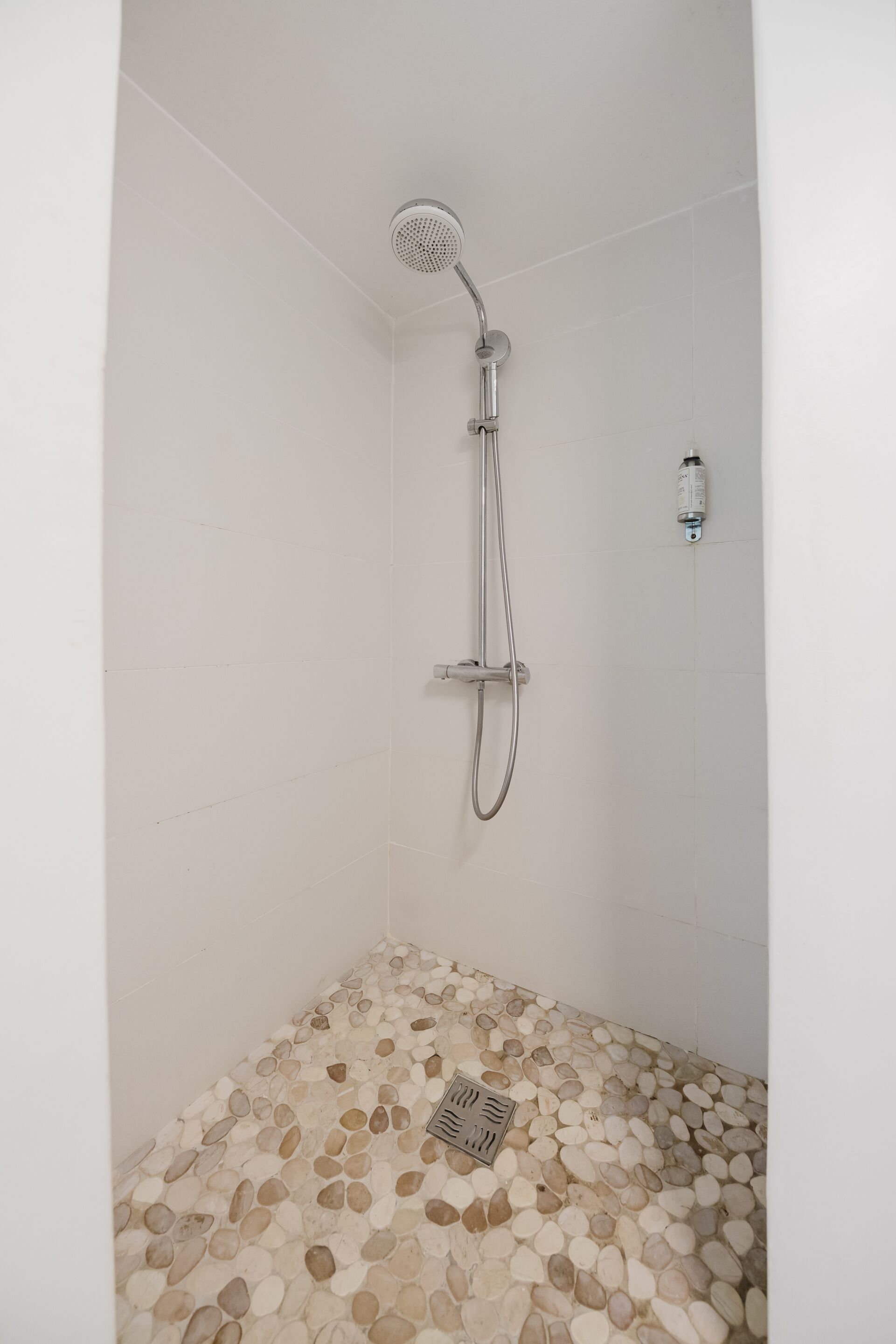 Ducha con cabezal de ducha de mano y controles fijos en la pared. El suelo de la ducha está cubierto de guijarros.