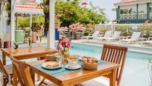 A wooden table with breakfast items, a poolside setting, and a view of a house with a red roof. - Hôtel Frégate Bleue (Le Francois)