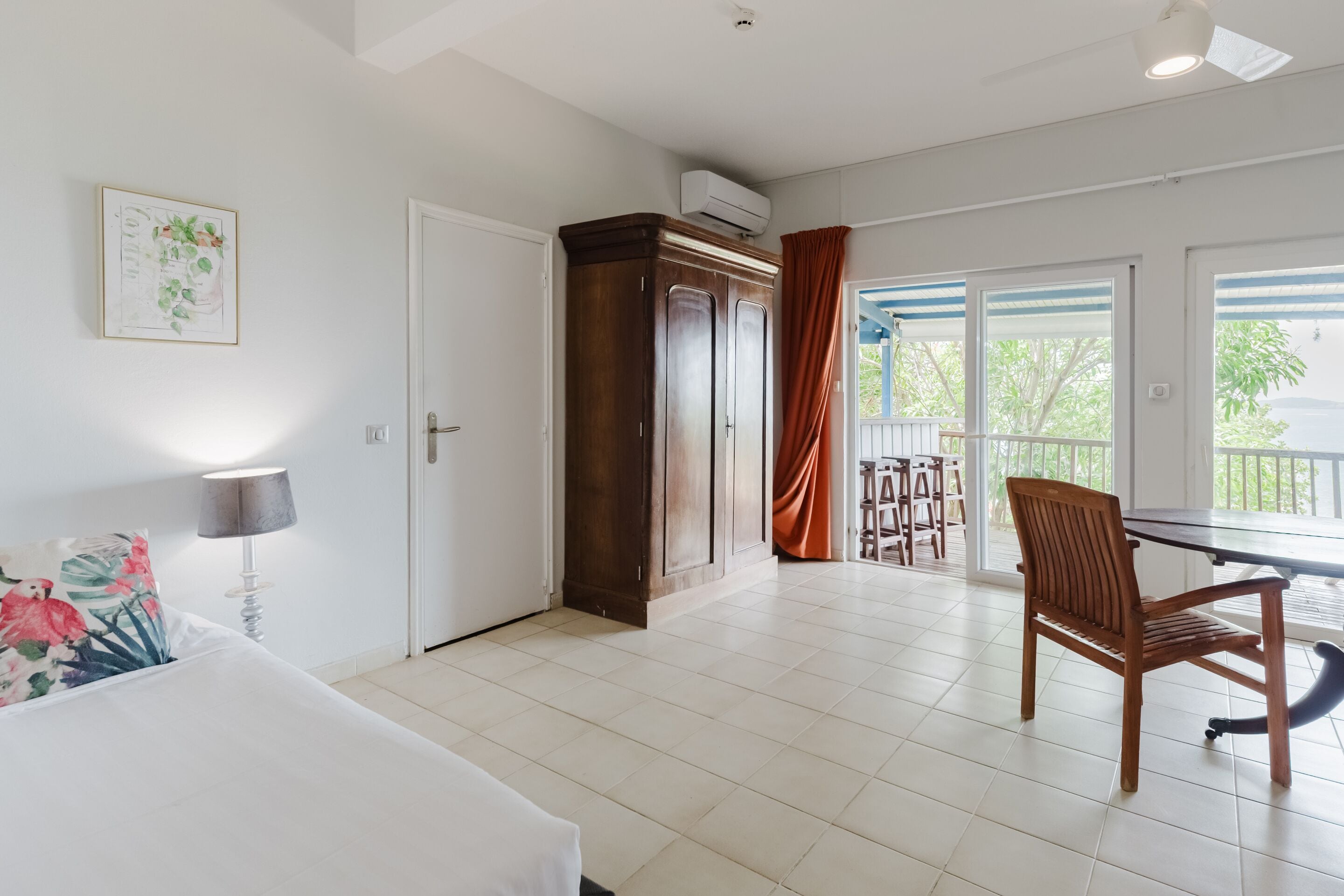 A hotel room with a bed, a wooden wardrobe, a dining table with chairs, and a balcony with a view.