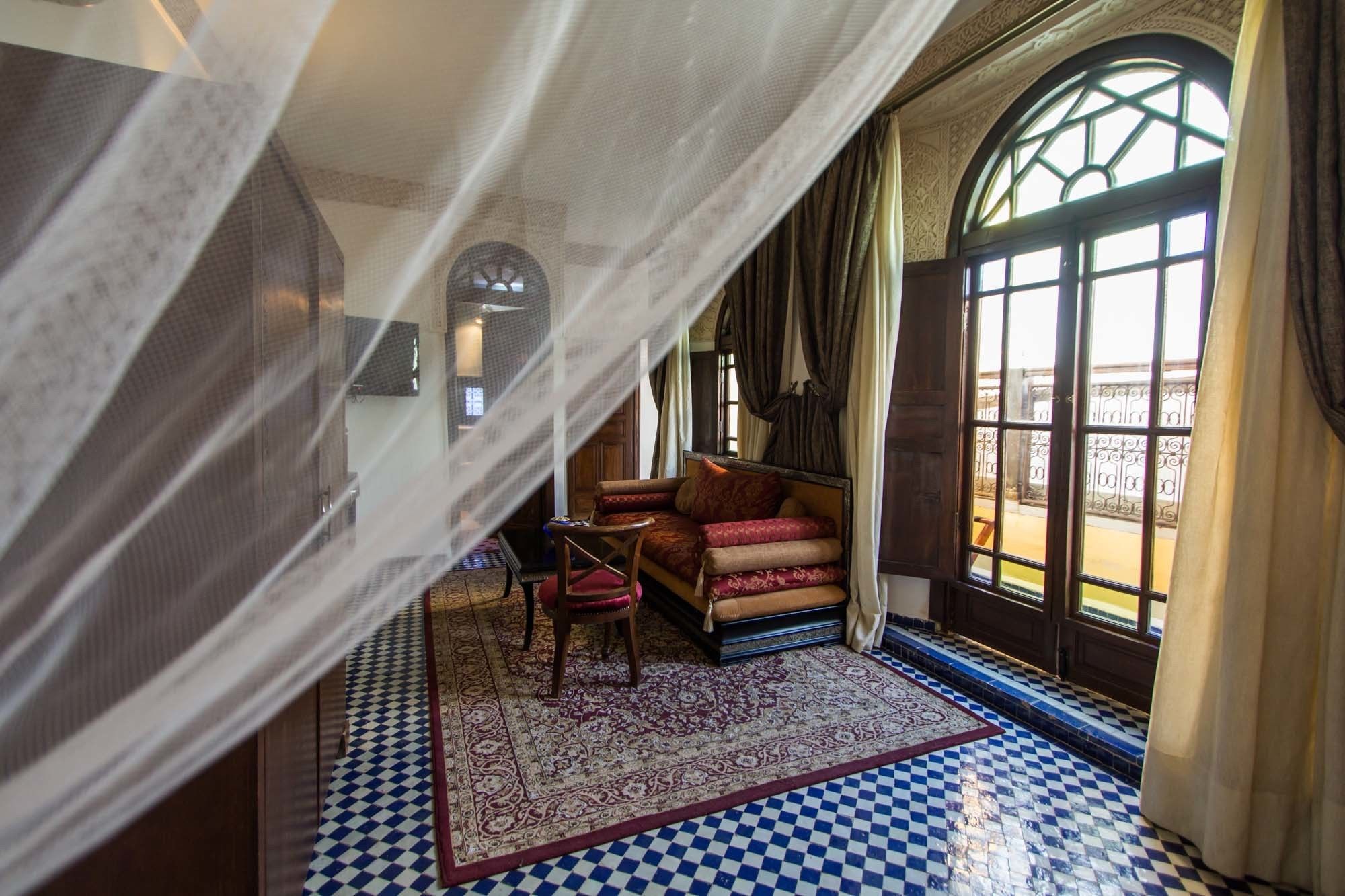 A room with a checkered floor, a sofa with patterned cushions, a chair, and a large window with curtains.
