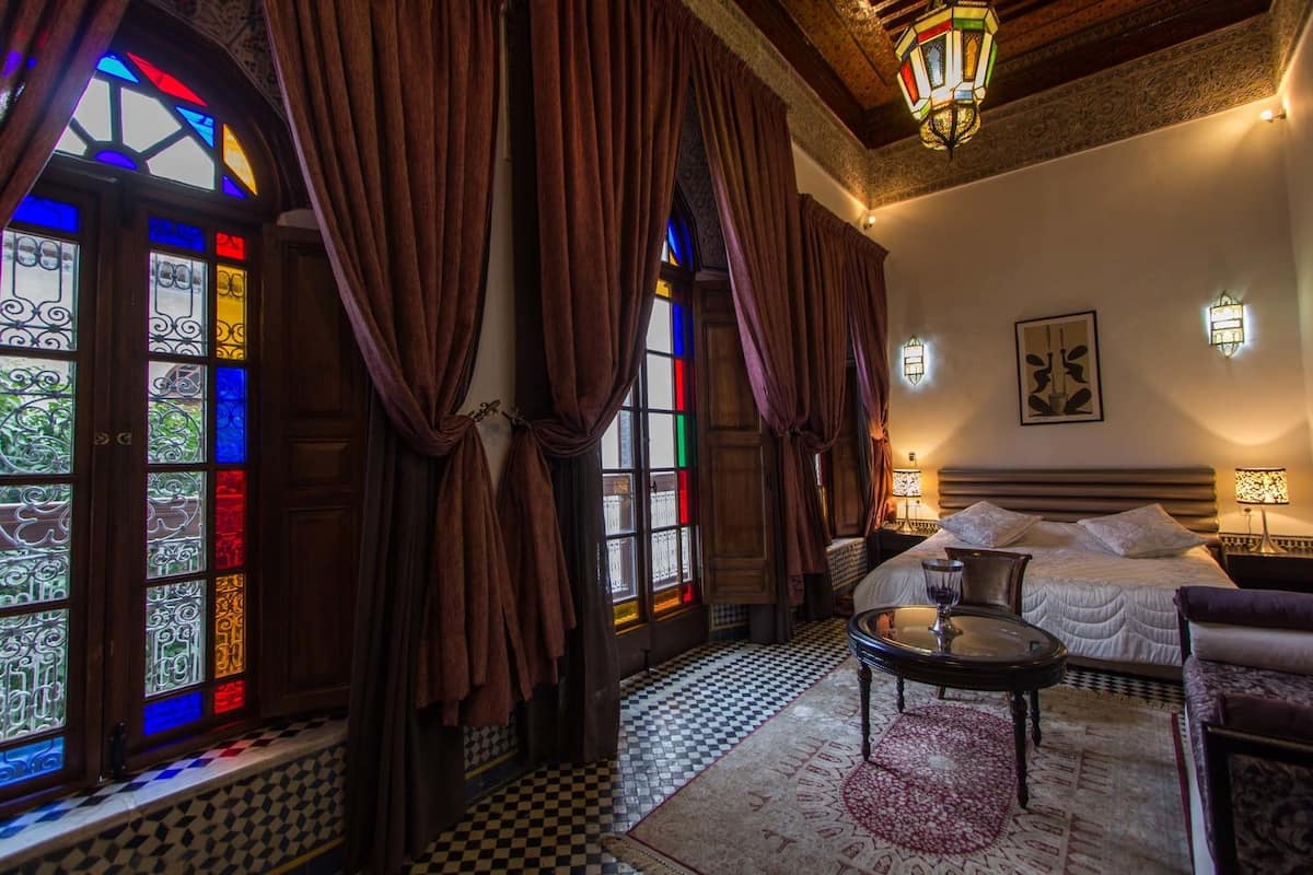 A room with a checkered floor, a bed with white linens, a small round table, and a chair.